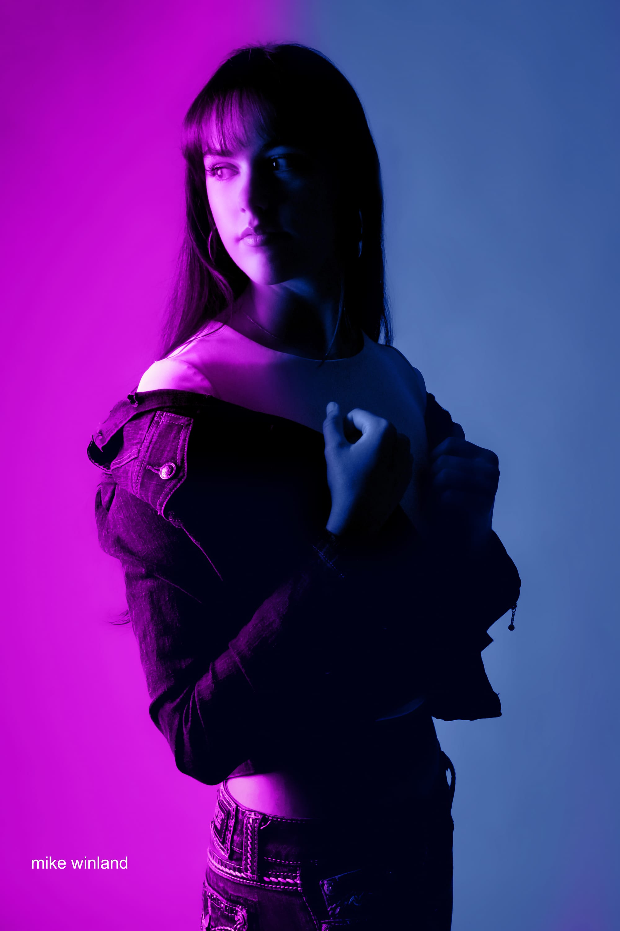 Senior portrait lit with magenta and blue gel light, the subject wearing a denim jacket off the shoulder.