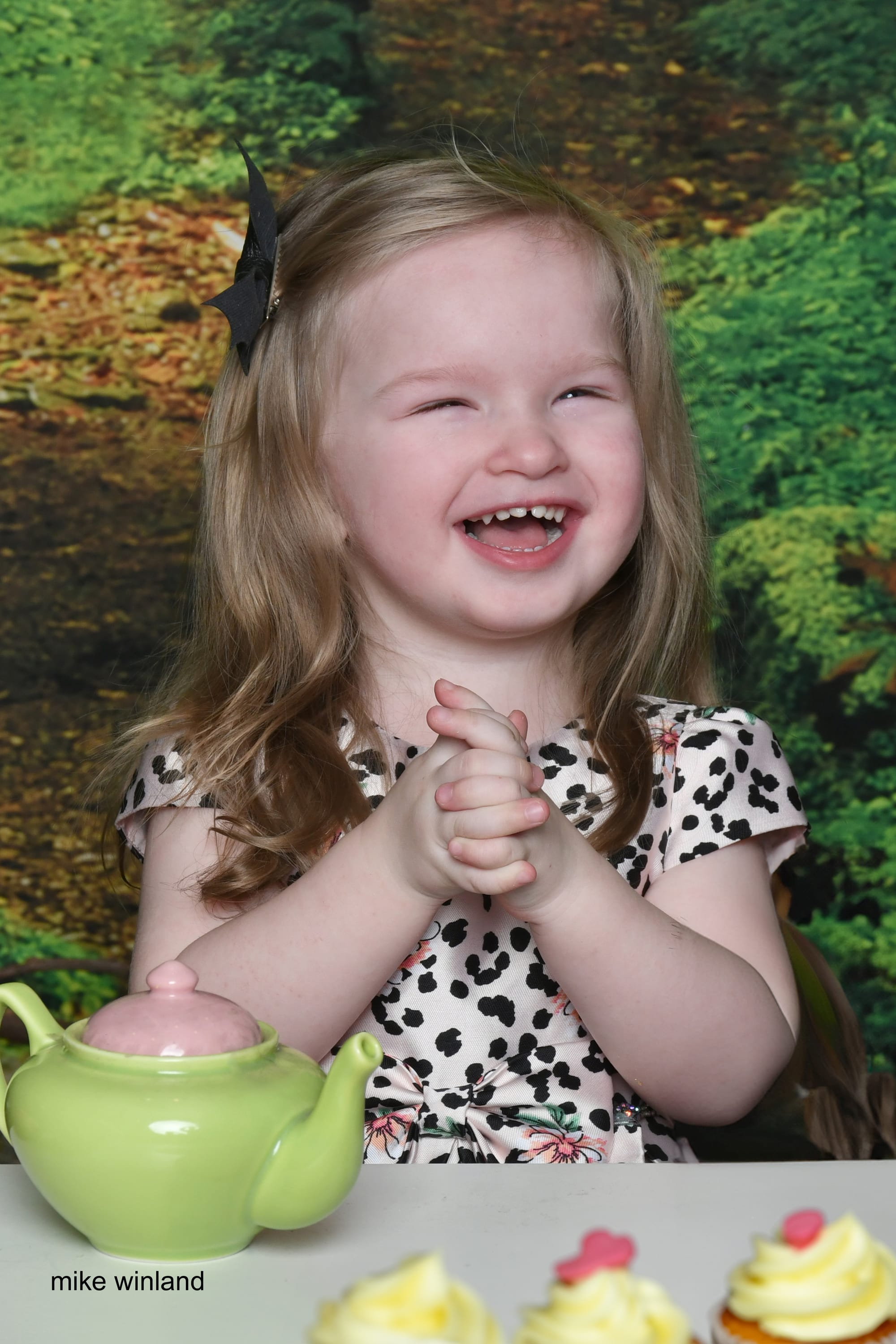 Young girl in a spotted dress laughing at a table set with a green teapot and frosted cupcakes in front of a woodland backdrop.